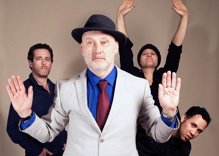 Photo of the band with four people in it, with Jah Wobble at the front holding this hands up.