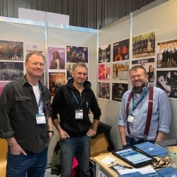 Barry Stewart, Pink Thomas and Matt Bartlett, stood in front of pictures of artists behind a trade stand at a trade fair.